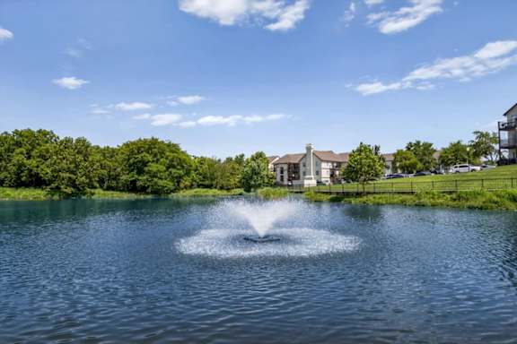 Waterfront lake with fountain surrounded by manicured grounds and residential buildings.