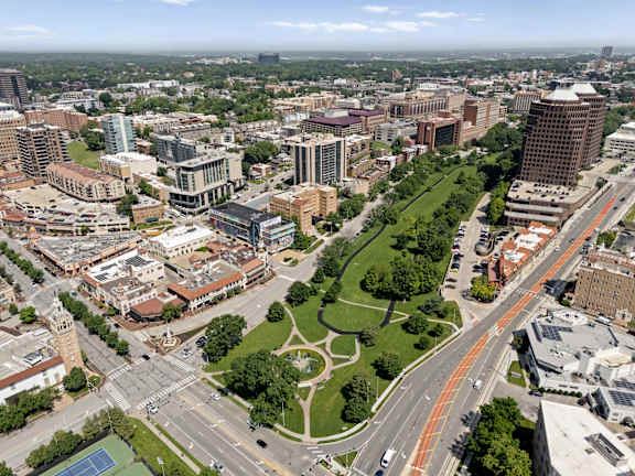 A cityscape with a mix of modern and older buildings, a large green park area, and a busy road intersection.