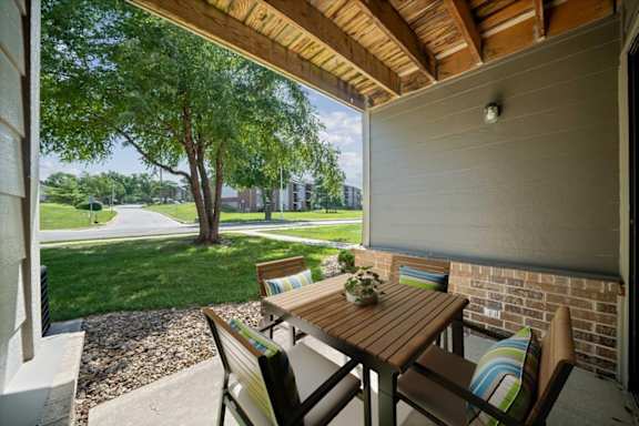 Private covered patio at Wind River Lodge Apartments featuring outdoor seating, landscaped grounds, mature trees, and views of the community.