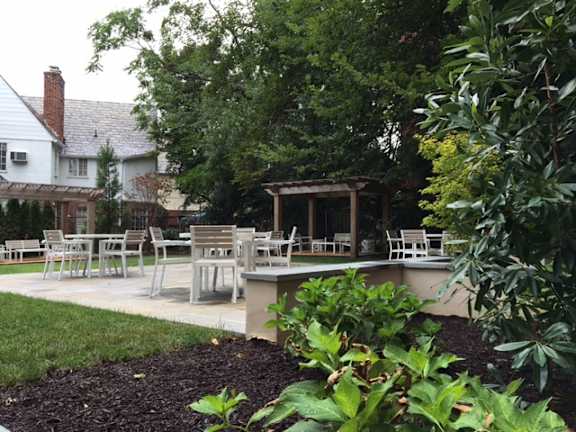 A patio with chairs and a table is surrounded by green plants.