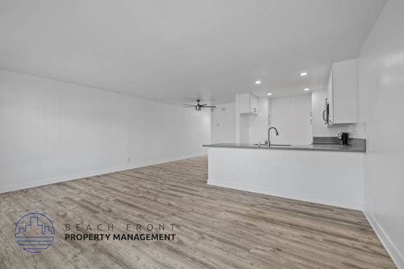 A spacious room with wooden flooring and white walls, featuring a kitchenette with a sink and a refrigerator.