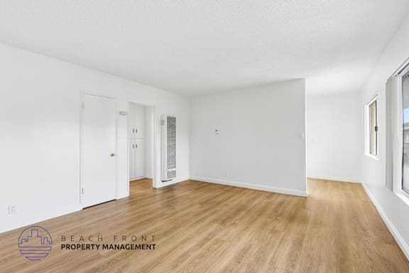 A spacious room with wooden flooring and white walls, with a door on the left and a window on the right.