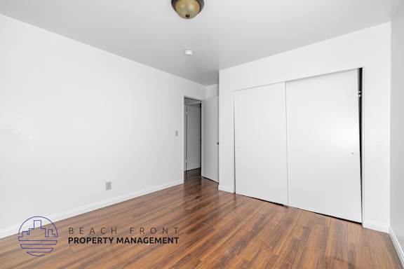 A room with a wooden floor and white walls, with a logo for Beach Front Property Management in the bottom left corner.