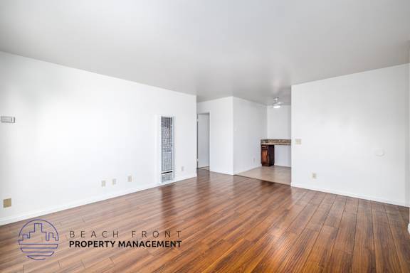 A spacious room with wooden flooring and white walls, with a logo for Beach Front Property Management in the bottom left corner.