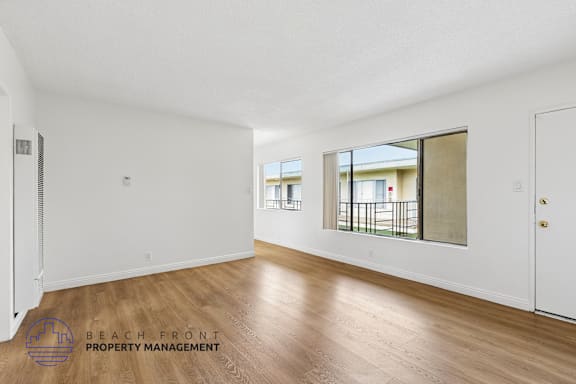 A spacious room with wooden flooring and a view of a balcony through a window.