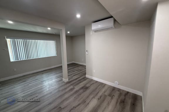 A room with a window covered by blinds, wooden flooring, and a wall-mounted air conditioner unit.