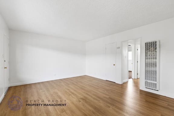 A spacious room with wooden flooring and white walls, with a logo for Beach Front Property Management in the bottom left corner.