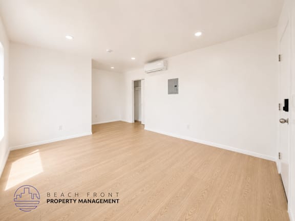 A spacious room with wooden flooring and white walls, with a logo for Beach Front Property Management in the bottom left corner.