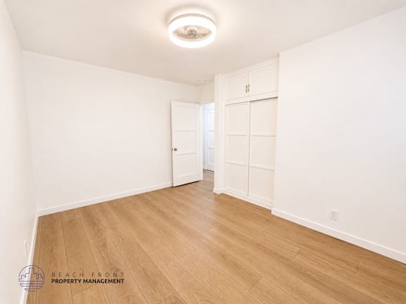 A spacious room with wooden flooring and white walls, with a logo for Beach Front Property Management in the bottom left corner.