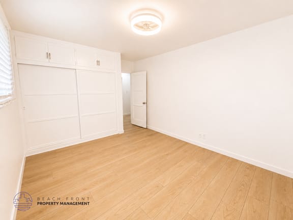 A white room with wooden flooring and a logo for Beach Front Property Management in the bottom left corner.