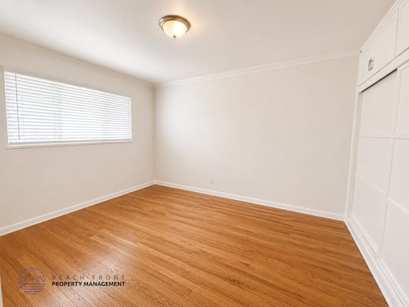 A room with wooden flooring and white walls, with a window on the left side.