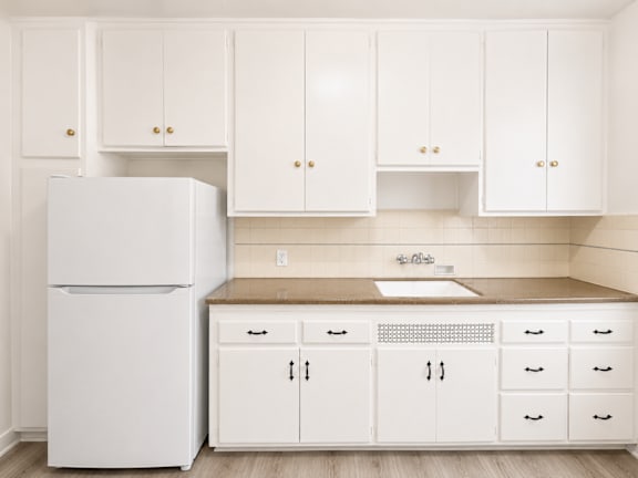 A white refrigerator stands next to a kitchen counter with white cabinets.