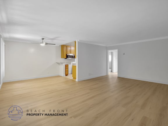 A spacious room with wooden flooring and white walls, featuring a kitchenette with wooden cabinets and a ceiling fan.