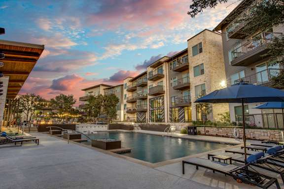 A swimming pool surrounded by lounge chairs and umbrellas in front of a modern apartment building.