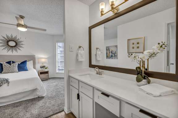 A well-lit, white bathroom with a large mirror and a bed in the background.