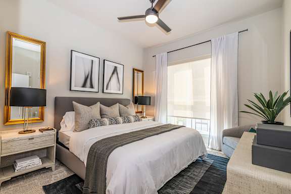 A well-lit bedroom with a large bed, a ceiling fan, and a potted plant.