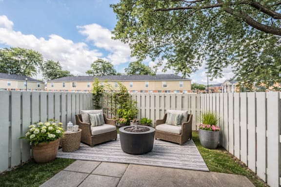 A white fence encloses a cozy outdoor seating area with a couch, chairs, and potted plants.