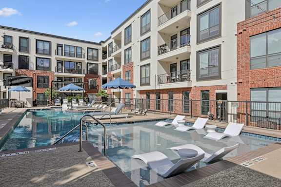 A large pool surrounded by lounge chairs and umbrellas in front of apartment buildings.