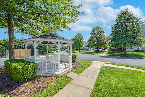 A white gazebo with a black roof is surrounded by greenery and a sidewalk at Acclaim at Sterling in Sterling, VA