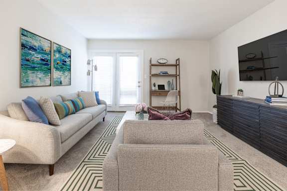 A living room with a beige couch, a grey ottoman, and a painting on the wall at Gramercy Apartments, Carmel, IN 46032