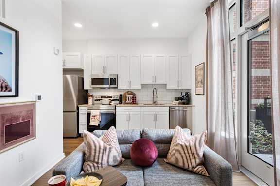A modern kitchen with white cabinets and a grey couch with a red pillow.