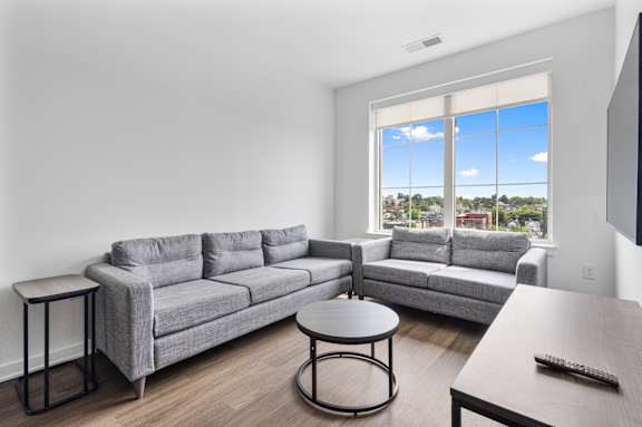 A grey couch sits in a living room with a large window showing a view of the city.