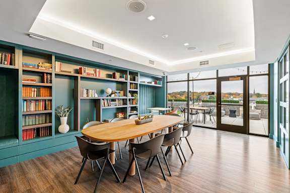 A room with a long table surrounded by chairs and bookshelves filled with books at Trio Newton Apartments, Apartments, Newton, MA