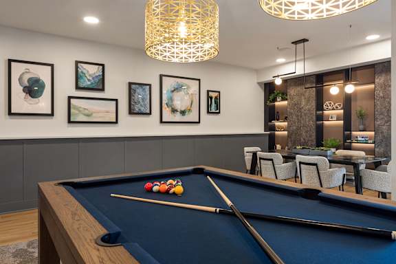 A pool table with a cue and balls is in the foreground of a room with a chandelier and framed pictures on the wall.