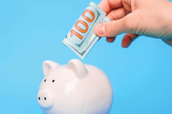 A hand is putting a 100 dollar bill into a piggy bank.