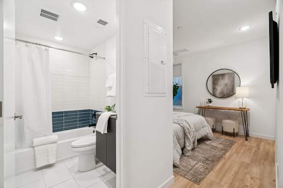 A bathroom with a white tub and toilet, a bedroom with a bed and desk, and a living area with a couch and a TV.