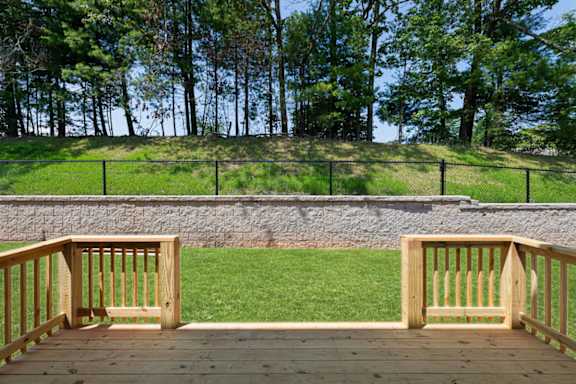 A wooden deck overlooks a grassy area with a stone wall and trees in the background.