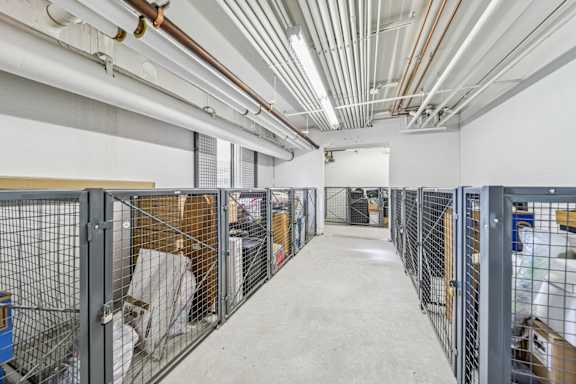 A long, narrow storage room with metal fencing and pipes on the ceiling.