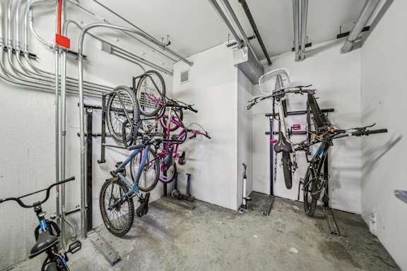 A bike rack with several bicycles attached is in a white room with pipes on the wall.