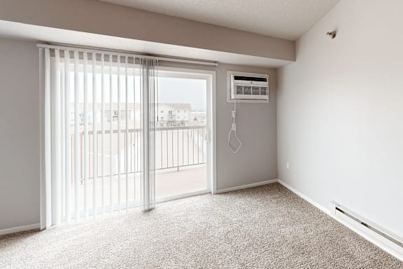 A room with a carpeted floor, a window with blinds, and a wall-mounted air conditioner unit.