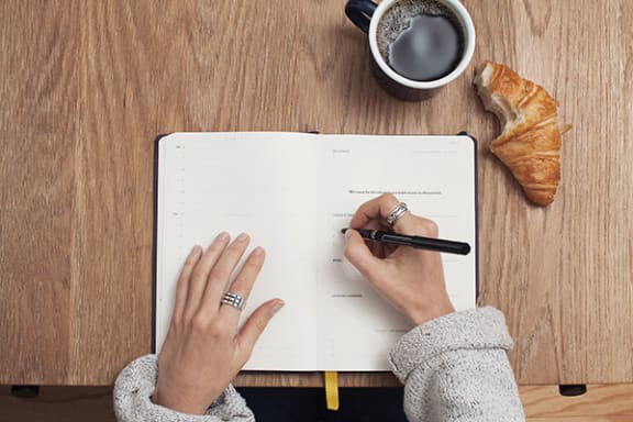 a woman writes in a notebook with a cup of coffee and croissant on the side