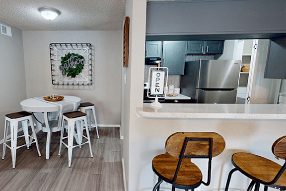 A kitchen with a table and chairs in the foreground and a fridge with an open sign in the background.