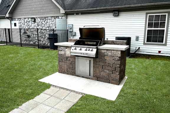 A stone and brick outdoor grill is on a patio in front of a house.