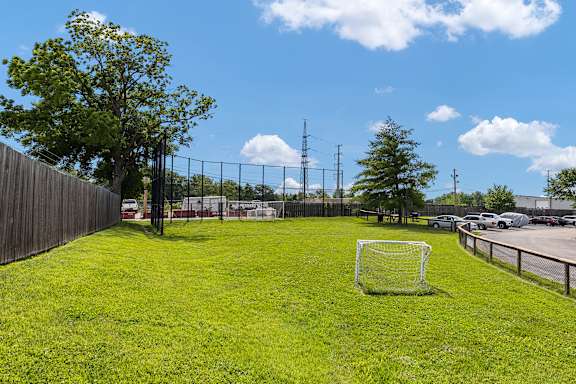 A soccer goal stands in a grassy field with a fence and trees in the background.