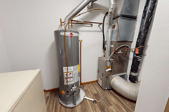 A water heater is installed in a room with a white wall and a wooden floor.