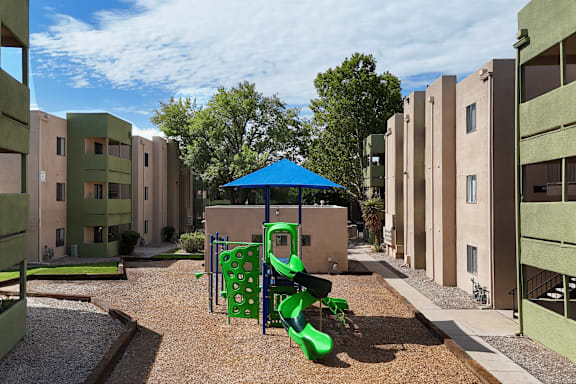 A playground with a blue umbrella and green slide in front of apartment buildings.