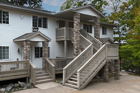 A house with a grey and white exterior has a wooden deck and stairs leading to a balcony.