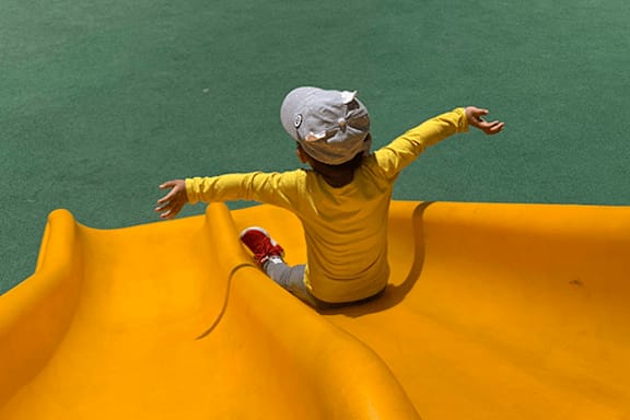 A child in a yellow shirt and red shoes is sliding down a yellow slide.