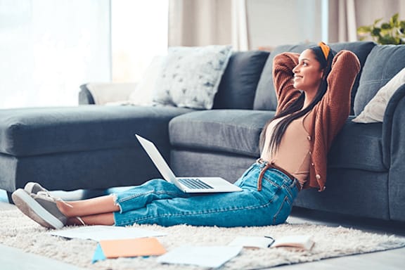 A woman is sitting on the floor with her laptop and her feet up on a couch.