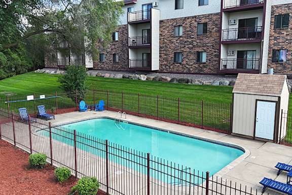 A pool surrounded by a black fence with blue chairs and a small building.