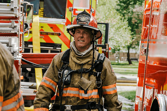 A firefighter in full gear stands in front of a fire truck.