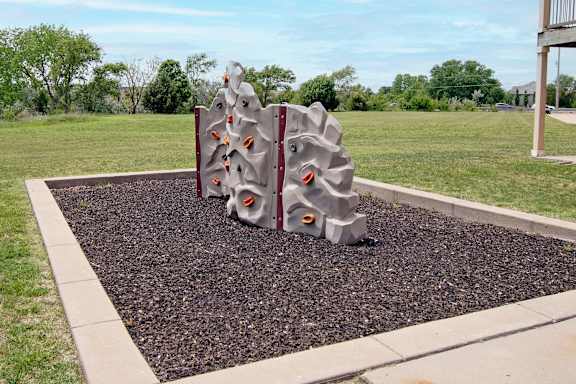 A playground structure with a grey and red color scheme is situated in a gravel area with a grassy background.