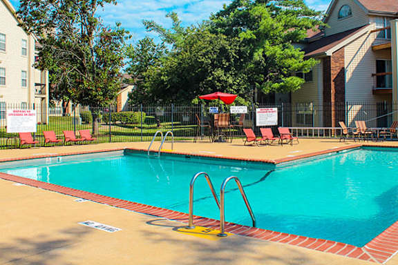 A pool with a red umbrella and chairs is surrounded by a fence.