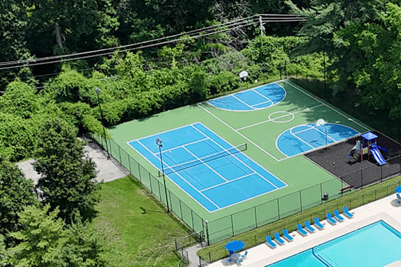 A tennis court is surrounded by trees and has a pool and lounge chairs.