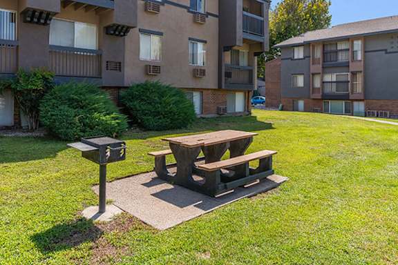 A wooden picnic table is situated in a grassy area in front of apartment buildings.