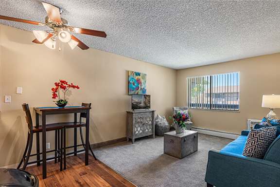A room with a ceiling fan, a dining table with chairs, a side table with a lamp, and a bed with a blue cover.